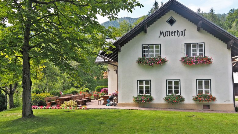 Ein traditionelles Bauernhaus mit dem Namen Mitterhof, umgeben von gr&uuml;ner Landschaft und Blumenbeeten.