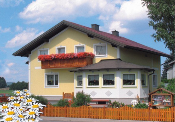 Ein gelbes Haus mit rotem Dach und Blumenkasten auf dem Balkon, umgeben von einem Holzzaun und G&auml;nsebl&uuml;mchen im Vordergrund.