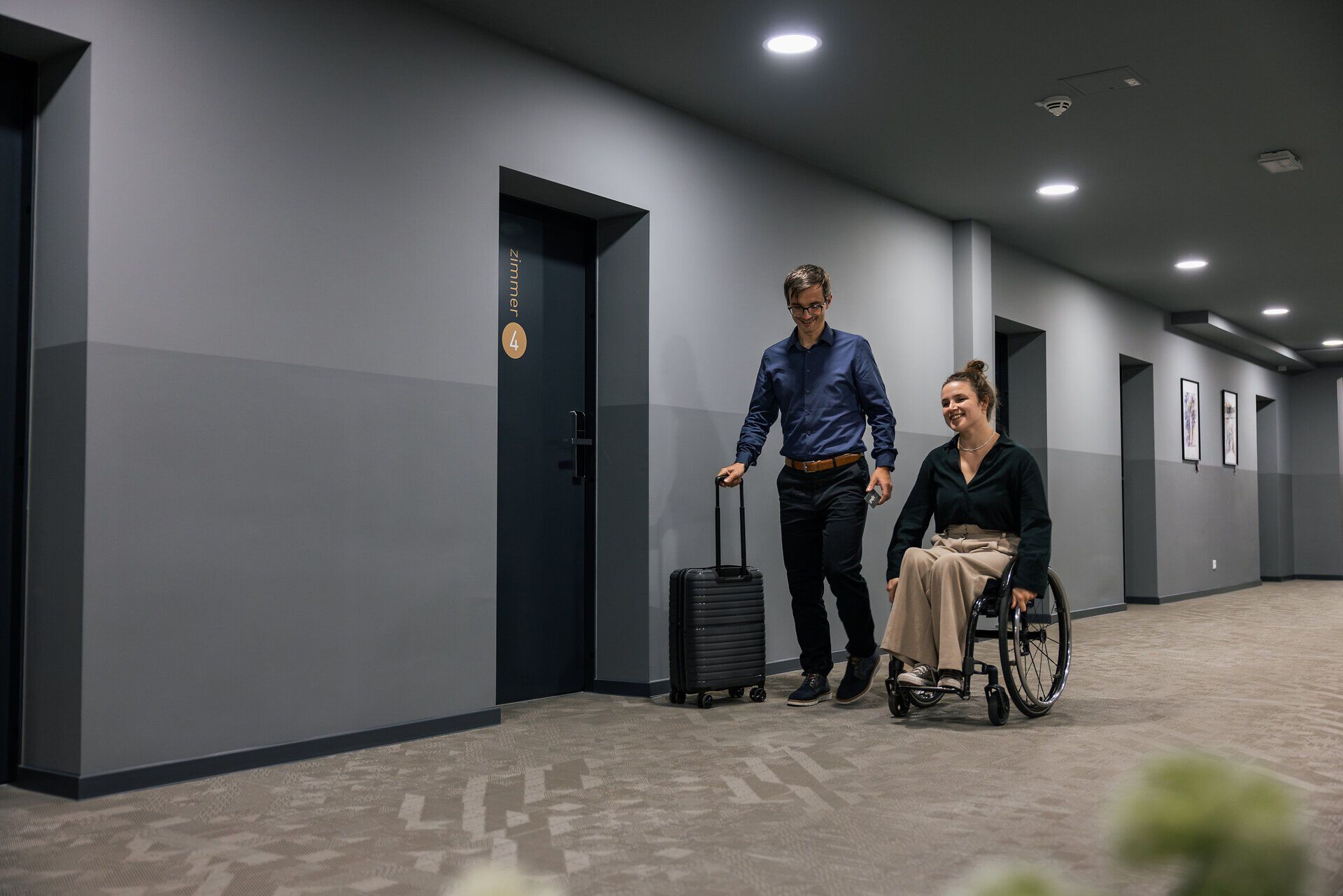 Eine junge Frau im Rollstuhl und ein Mann mit Koffer befinden sich auf dem Hotelgang am Weg zu ihrem Hotelzimmer.