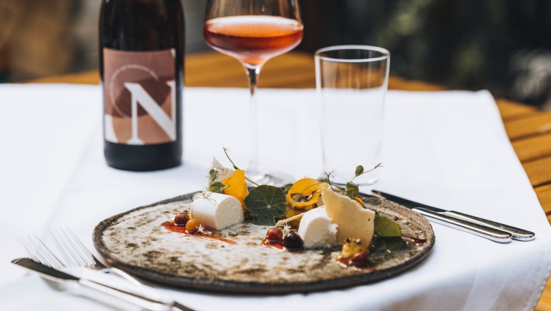 Ein Teller mit K&auml;se und Garnitur, ein Glas Ros&eacute;wein und eine Weinflasche auf einem Tisch.