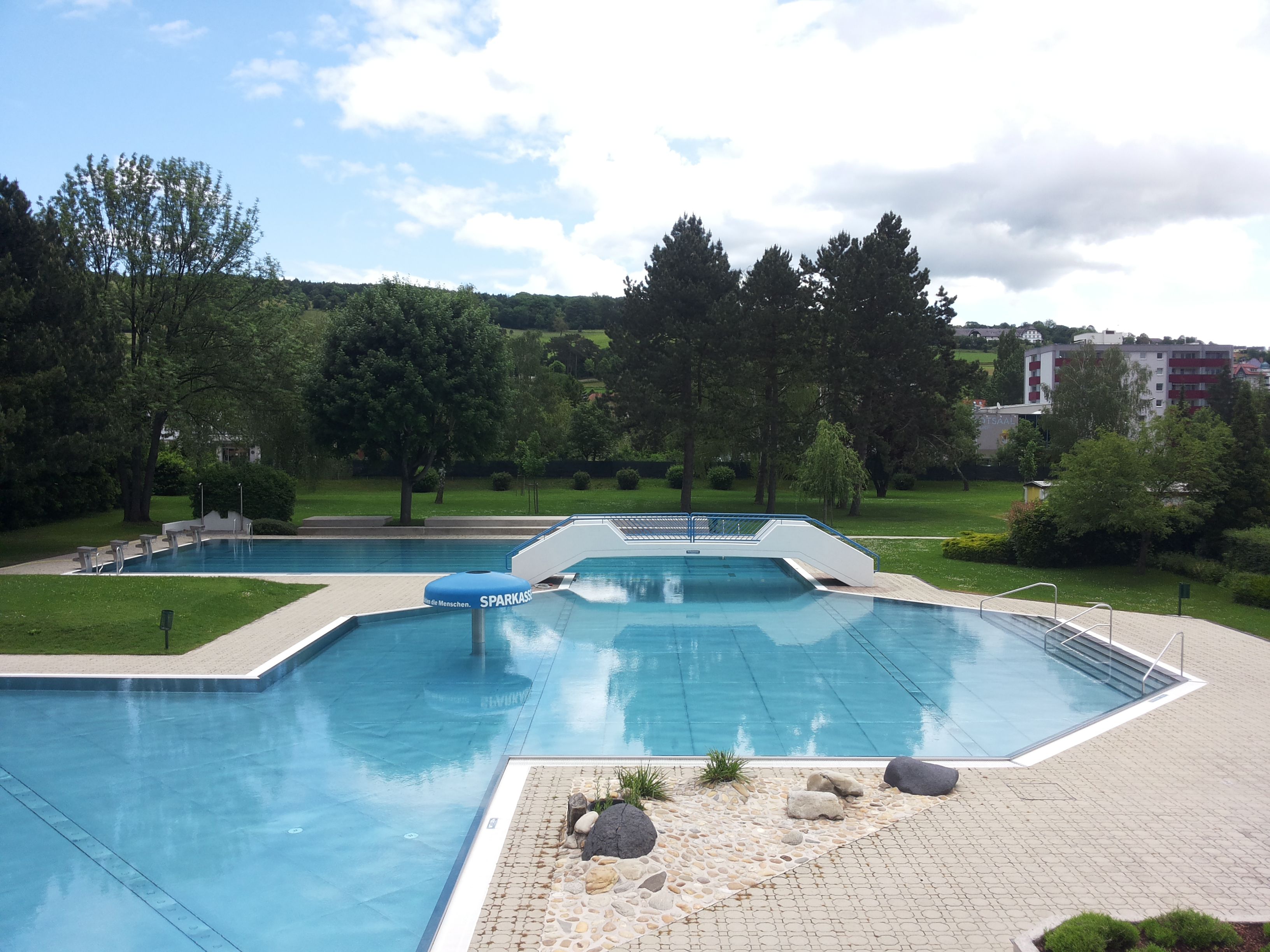 Ein leeres Freibad mit mehreren Becken, umgeben von Bäumen und Rasenflächen, unter einem bewölkten Himmel.