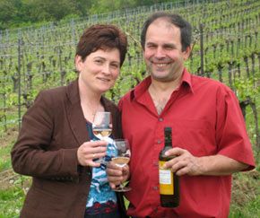 Ein Mann und eine Frau stehen in einem Weinberg und halten Weingläser und eine Weinflasche.