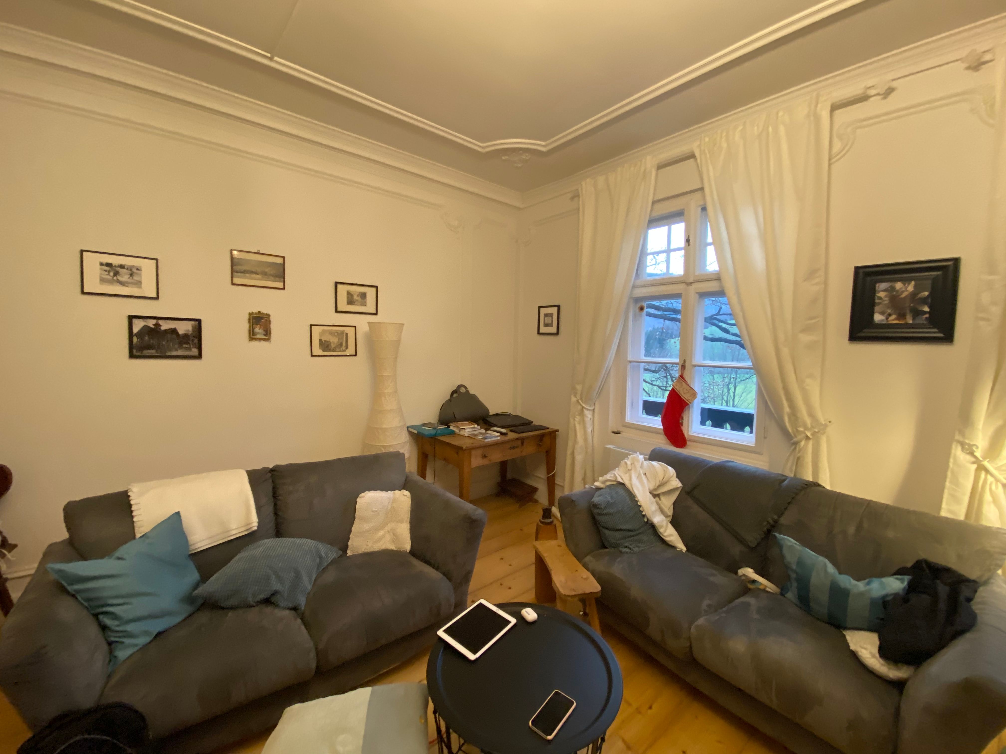 Gemütliches Wohnzimmer mit zwei grauen Sofas, einem runden Tisch, Bildern an der Wand und einem Fenster mit Vorhängen und Weihnachtsstrumpf.