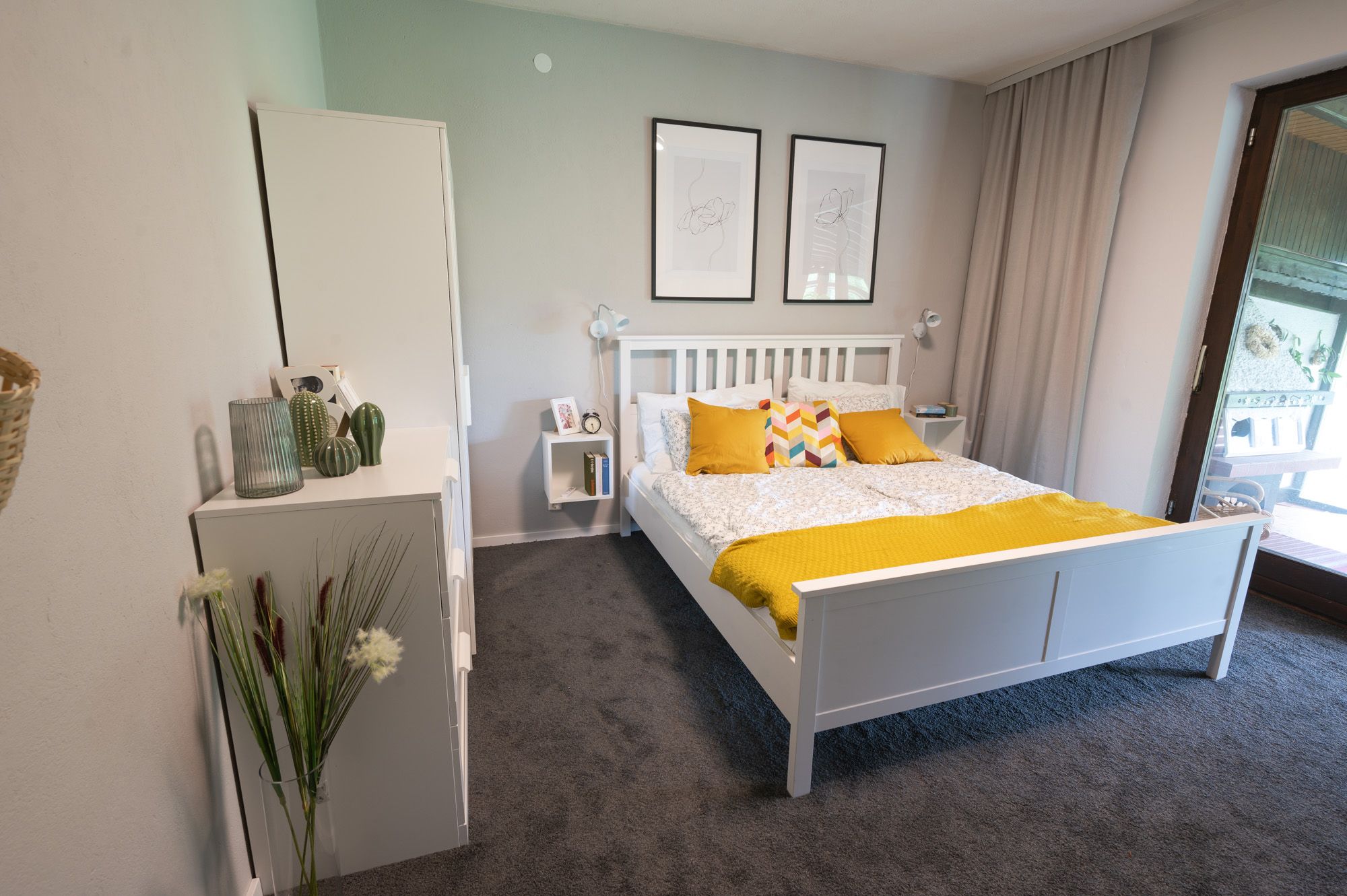 Schlafzimmer mit weißem Bett, gelben Kissen und Decke, zwei Wandbildern und einem Fenster mit Vorhang.