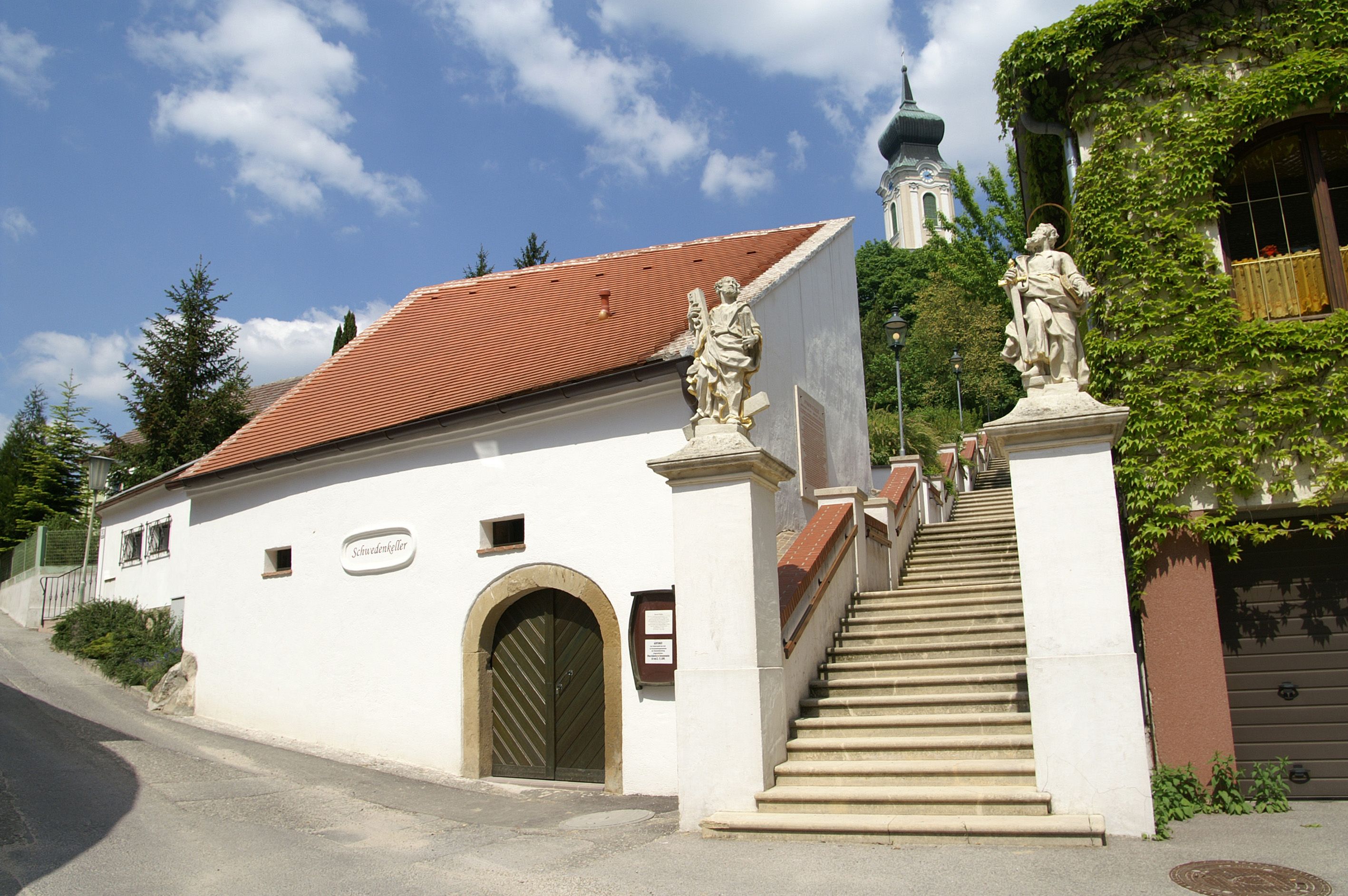 Weißes Gebäude mit rotem Dach und Treppe mit Statuen, Kirche im Hintergrund.