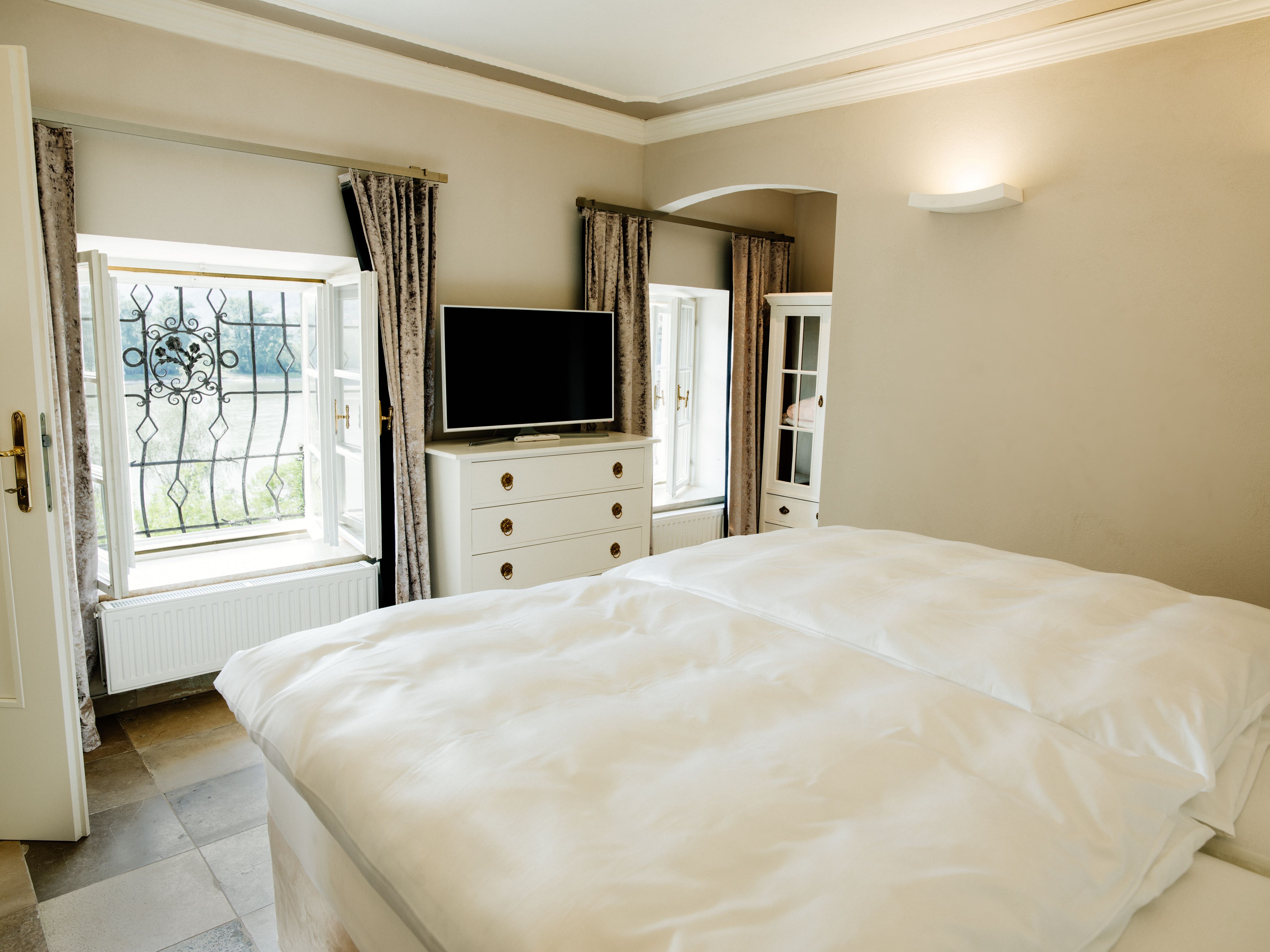 Helles Schlafzimmer mit Doppelbett, TV, Kommode und zwei Fenstern mit Vorhängen.