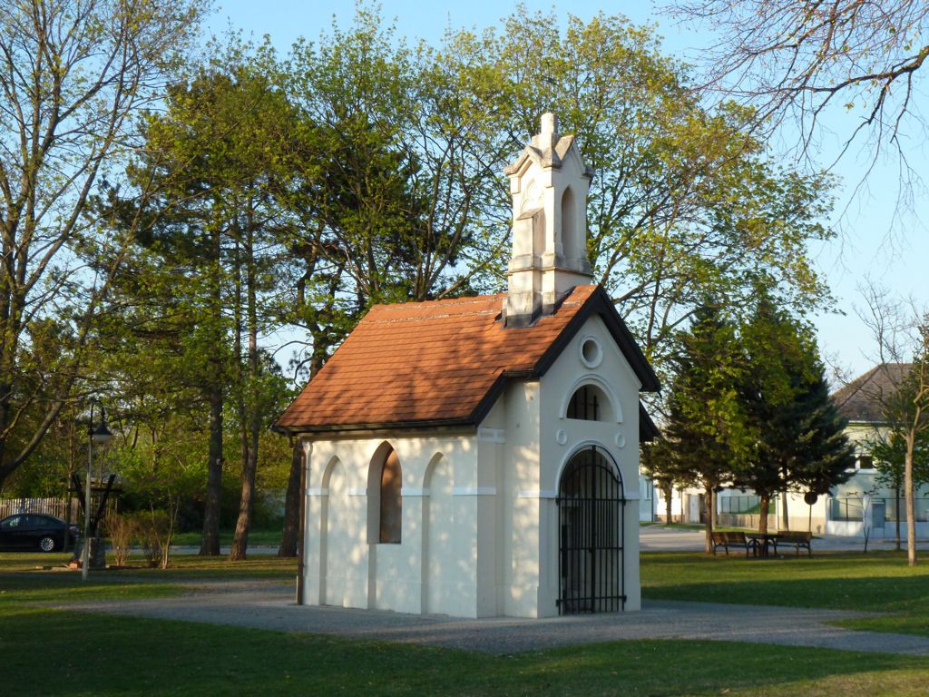 Eine kleine weiße Kapelle mit rotem Dach in einem Park, umgeben von Bäumen.