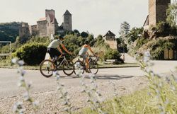 Zwei Radfahrer fahren entlang eine Straße, hinter ihnen die Burg Hardegg, im Vordergrund Blumen