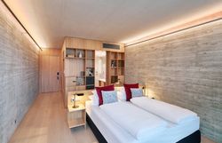 Ein hochmodernes Hotelzimmer in minimalistischem Design. Die linke Raumhälfte besteht aus hellem Naturholz, das nahtlos vom Boden in eine deckenhohe Regalwand und die Zimmertür übergeht. Rechts bildet eine Wand in Sichtbeton-Optik einen kühlen Kontrast. Das Doppelbett im Vordergrund ist schlicht weiß bezogen und wird durch zwei weinrote sowie zwei gemusterte Zierkissen akzentuiert. Eine dezente, indirekte Beleuchtung an der Decke sorgt für eine warme Atmosphäre.
