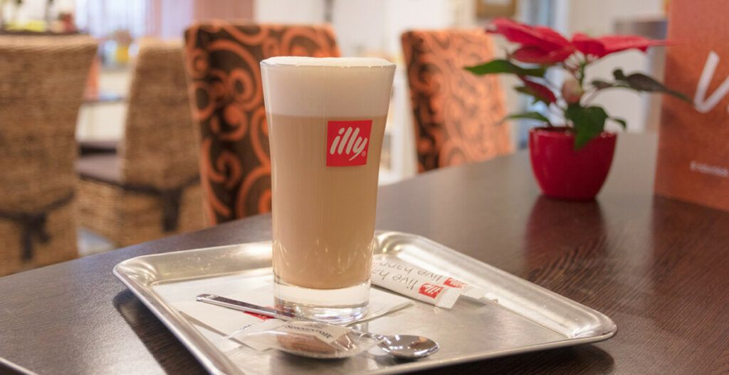 Ein Glas Café Latte auf einem Tablett mit einem Löffel und Zucker, im Hintergrund ein roter Blumentopf auf einem Tisch.