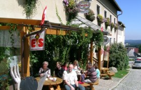 Eine Gruppe von Menschen sitzt vor einem Gasthof an Holztischen auf einer gepflasterten Straße.