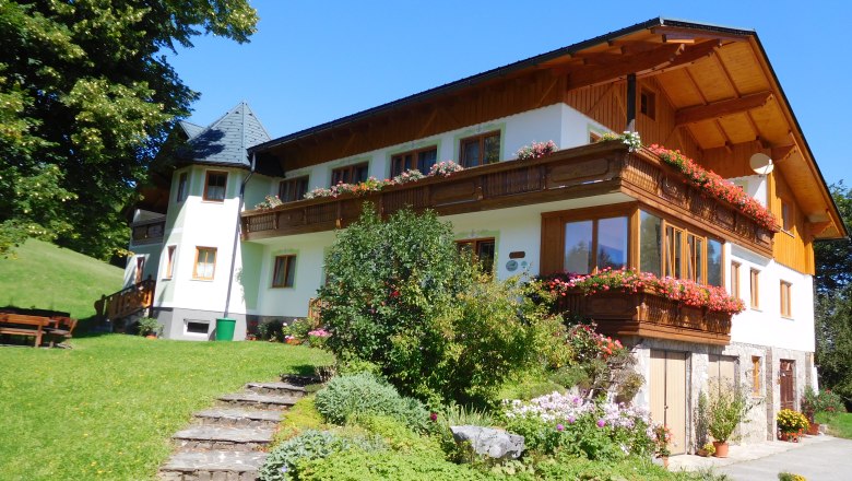 Ein traditionelles Bauernhaus mit Holzbalkon und bl&uuml;henden Blumen, umgeben von gr&uuml;ner Landschaft und blauem Himmel.
