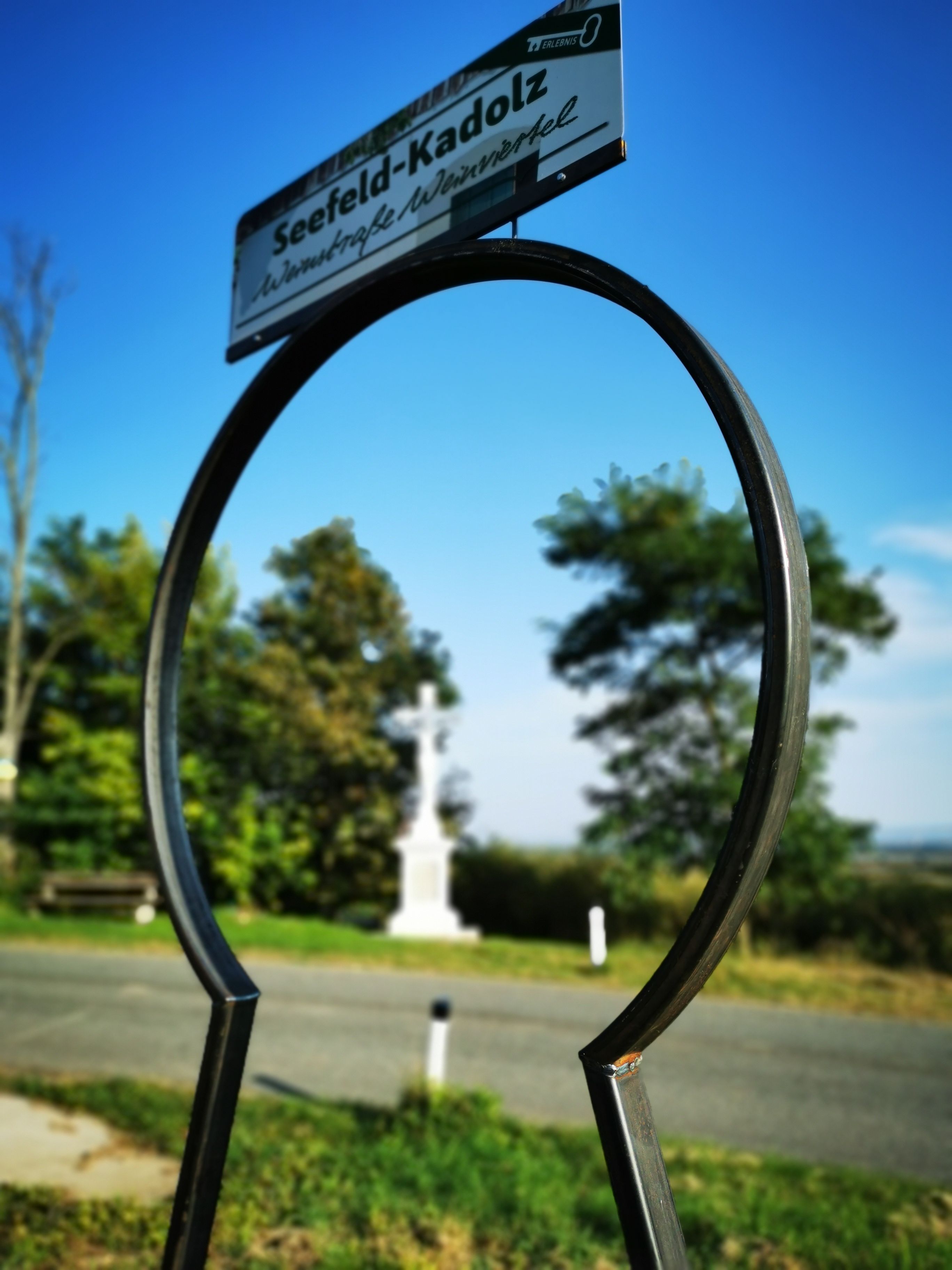 Ein Schild mit der Aufschrift 'Seefeld-Kadolz' vor einem blauen Himmel, im Hintergrund ein weißes Kreuz und Bäume.