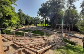 Ein Motorikpark mit verschiedenen Holzelementen und Kletterger&auml;ten, umgeben von B&auml;umen und Gr&uuml;nfl&auml;chen.