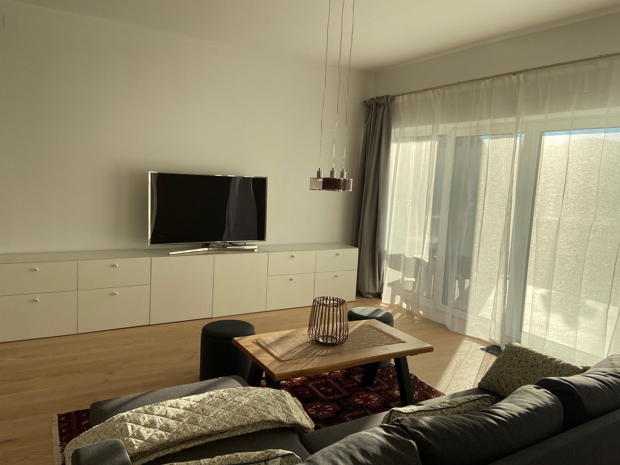 Moderner Wohnbereich mit grauem Sofa, Holztisch, Teppich, Fernseher und großen Fenstern mit Vorhängen.