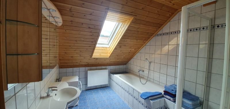 Ein Badezimmer mit Dachschräge, Dachfenster, Badewanne, Waschbecken und Dusche. Die Wände sind gefliest und die Decke ist aus Holz.