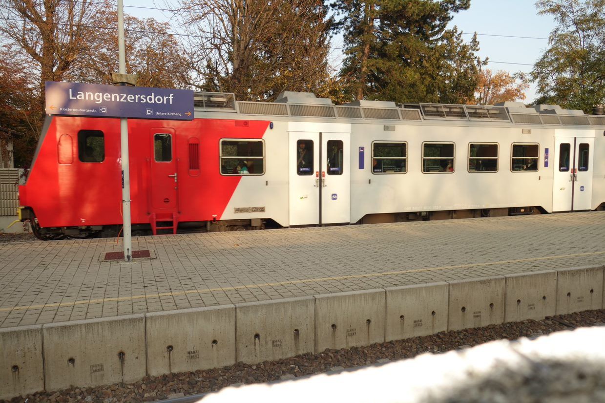 Ein Zug steht am Bahnhof Langenzersdorf, mit einem Schild im Vordergrund. Der Zug ist rot-weiß und die Umgebung ist von Bäumen umgeben.
