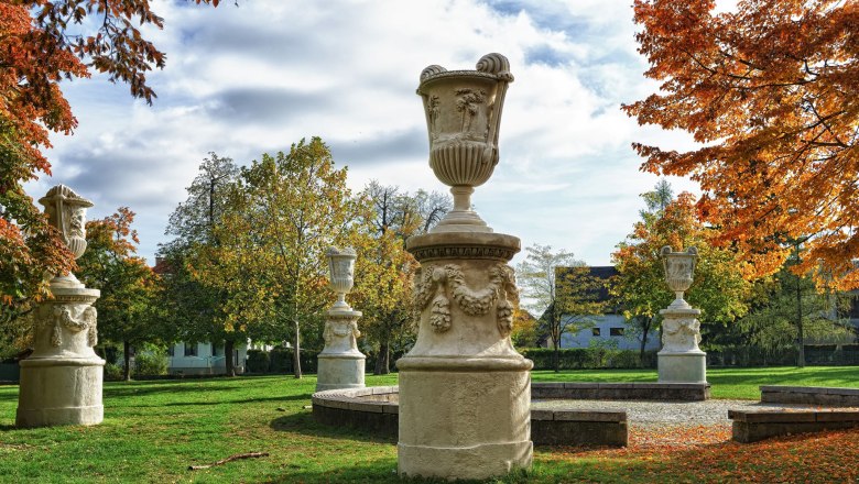 Steinskulpturen in einem herbstlichen Park mit buntem Laub und blauem Himmel.