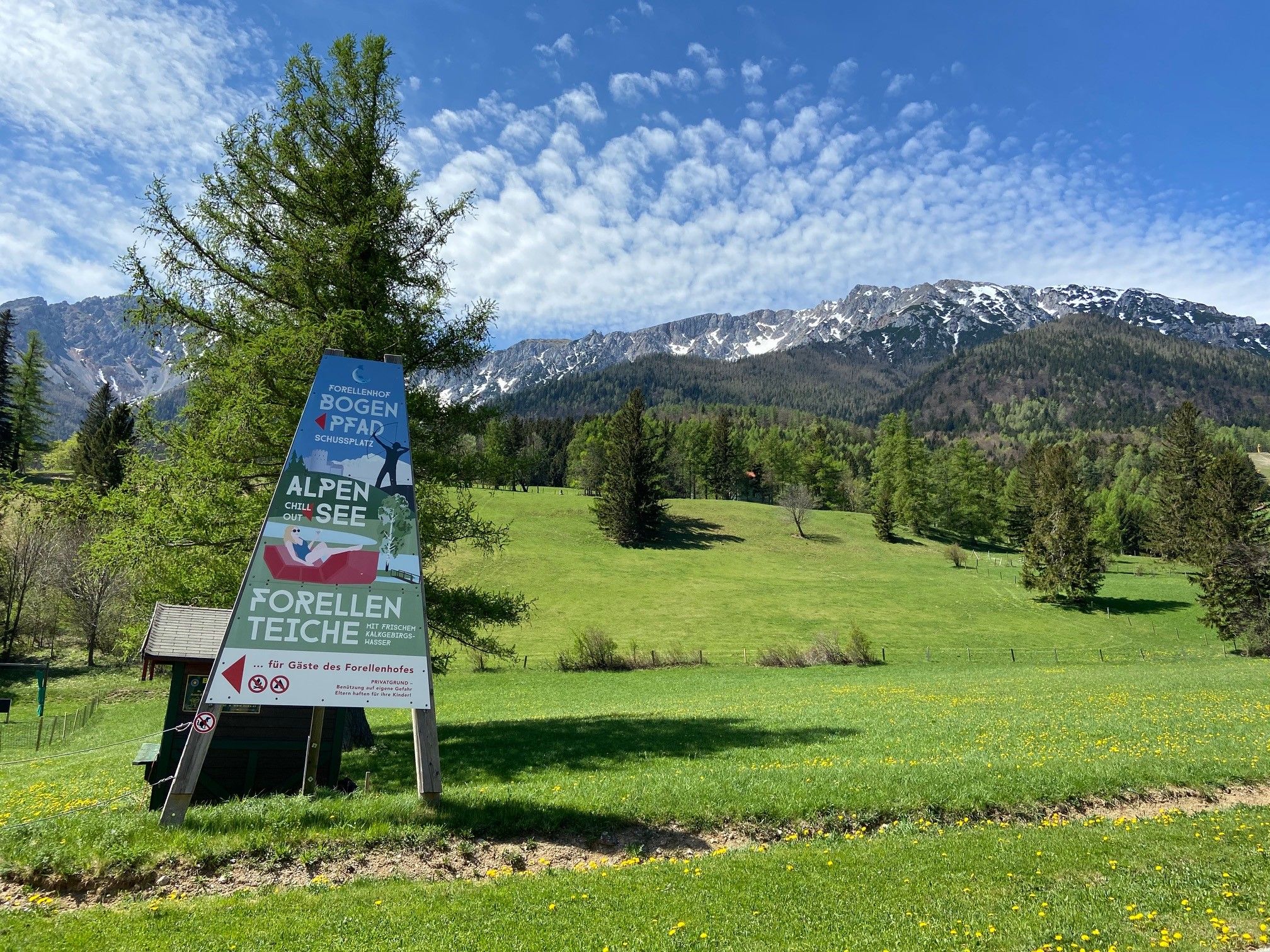 Schild vor Berglandschaft mit Wiese und Bäumen.