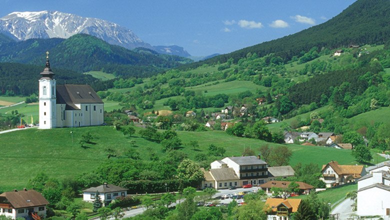 Landschaft mit Kirche auf H&uuml;gel, umgeben von gr&uuml;nen Feldern und Bergen im Hintergrund.