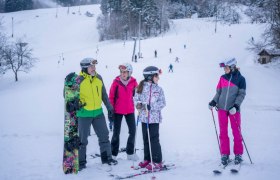 Gruppe von Skifahrern und einem Snowboarder auf einer verschneiten Piste mit Skilift im Hintergrund.