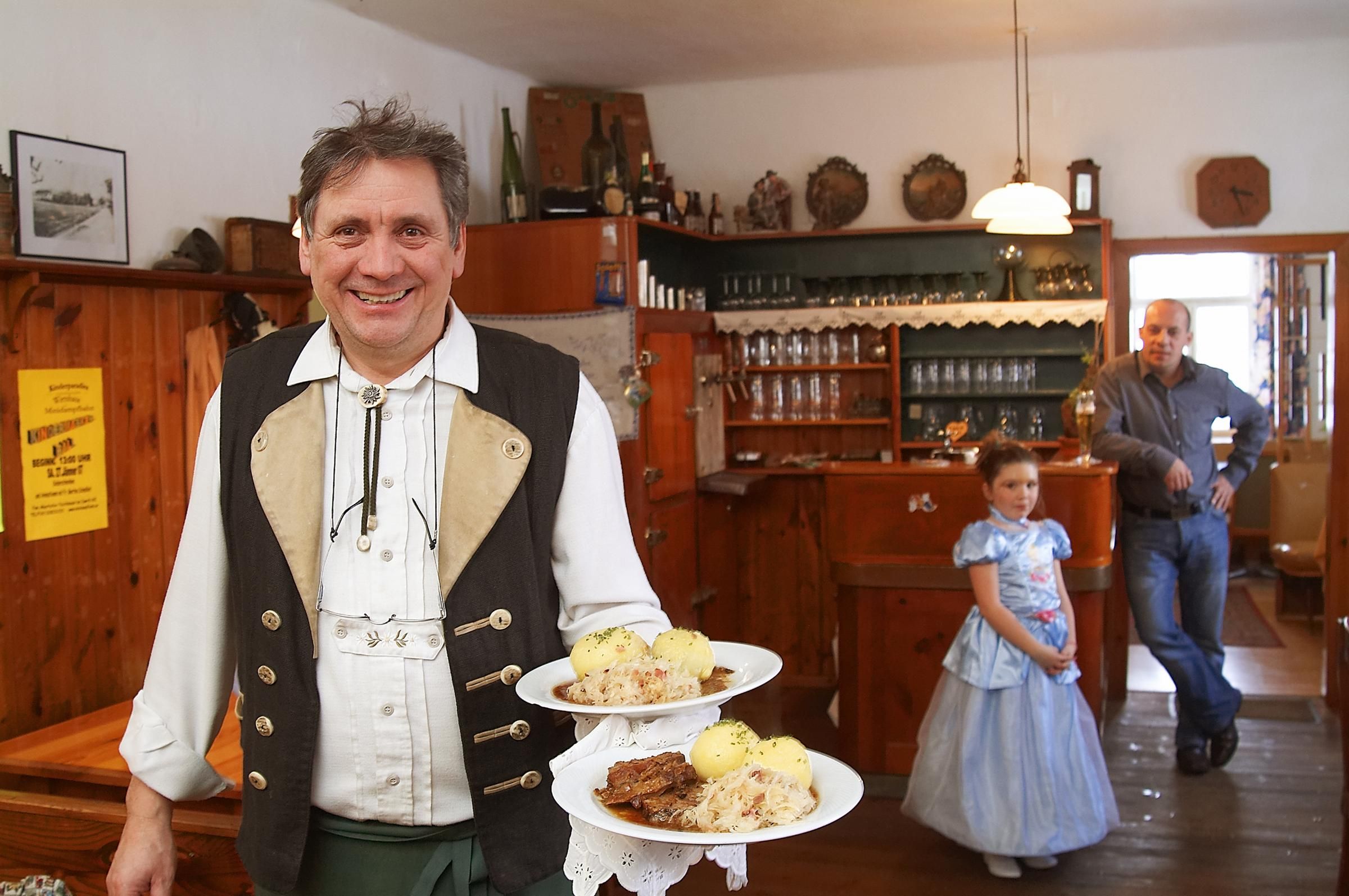 Ein Mann in traditioneller Kleidung serviert Schweinsbraten mit Knödeln in einem rustikalen Gasthaus. Im Hintergrund ein Mann und ein Mädchen im Kleid.
