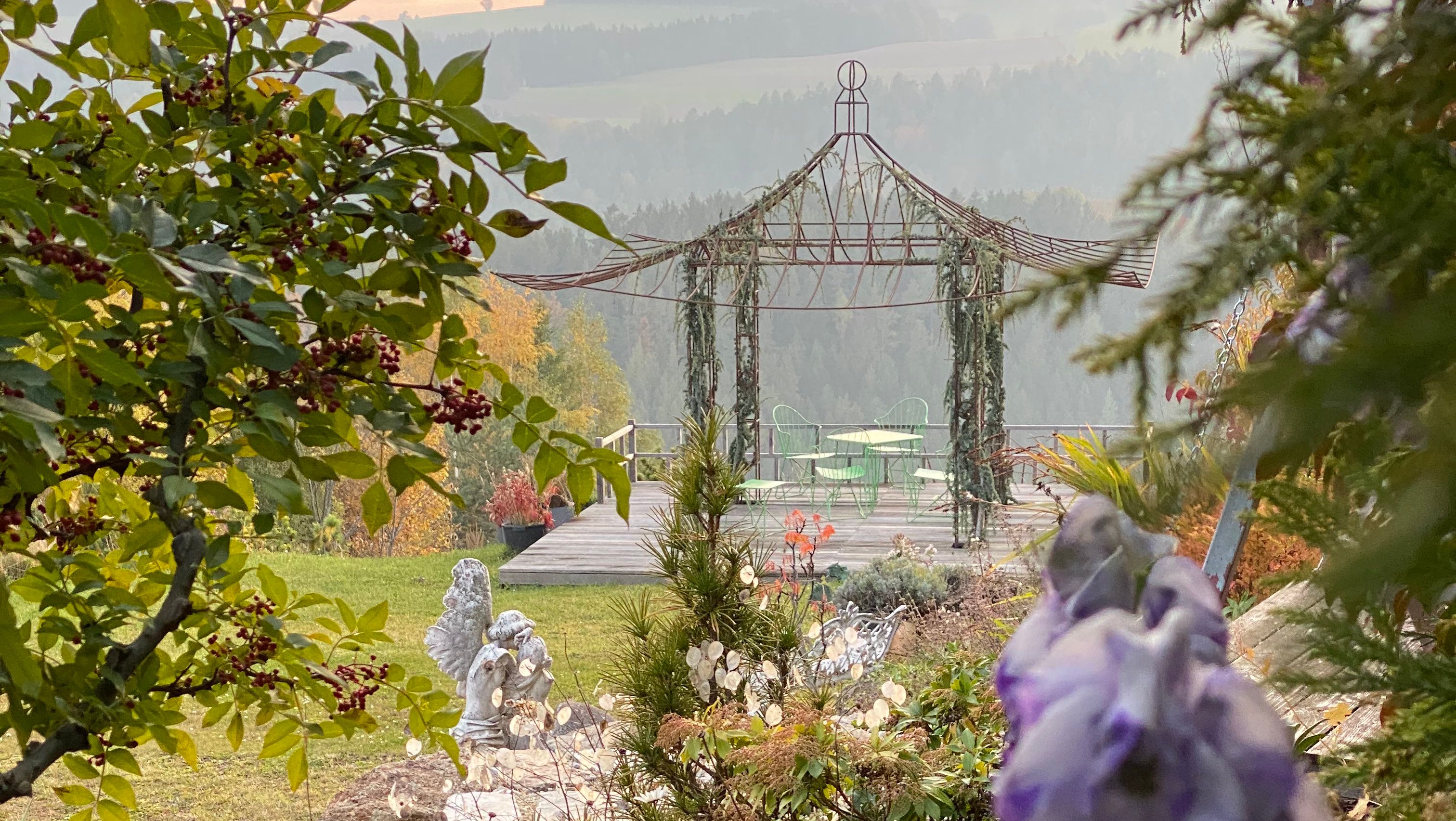 Ein romantischer Garten mit Pavillon, grünen Stühlen und Tisch, umgeben von Bäumen und Blumen.