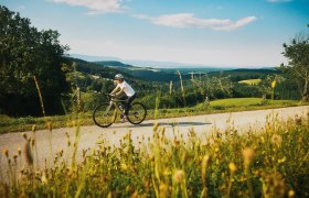 Eine Person f&auml;hrt mit einem Fahrrad auf einer l&auml;ndlichen Stra&szlig;e, im Hintergrund weiter Aussicht.