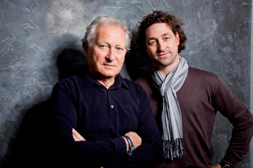 Zwei Männer stehen vor einer grauen Wand, einer älter mit verschränkten Armen, der andere jünger mit Schal.