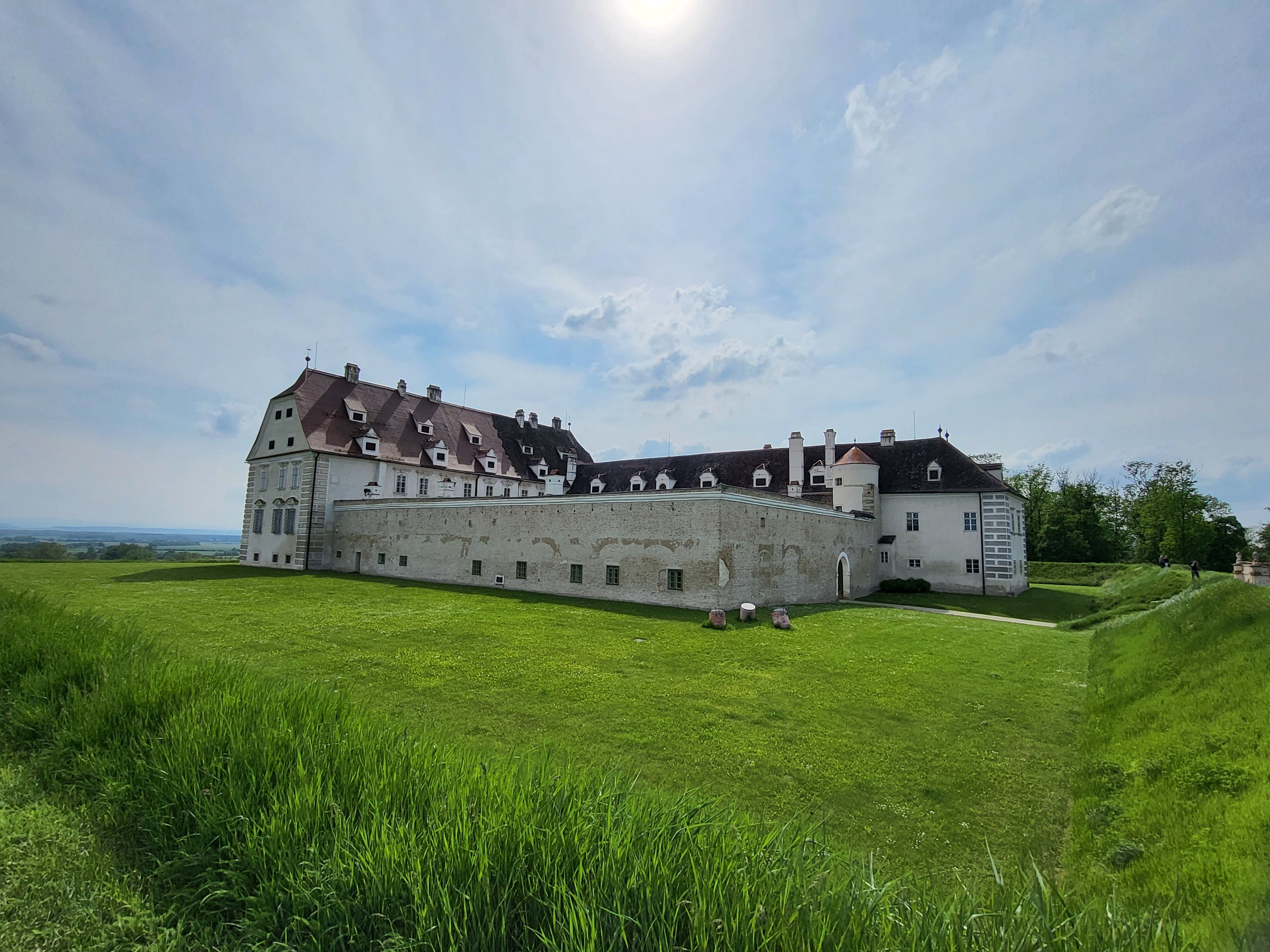 Ein großes, historisches Schloss mit weißen Mauern und roten Dächern steht auf einer grünen Wiese unter einem bewölkten Himmel.