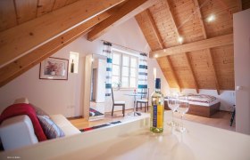 Gem&uuml;tliches Dachgeschosszimmer mit Holzbalkendecke, Bett, Sofa, Tisch und Weinflasche.