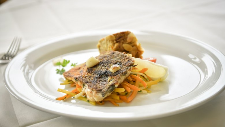 Ein Teller mit gebratenem Fischfilet auf Gem&uuml;sestreifen und einer Beilage auf wei&szlig;em Tischtuch.