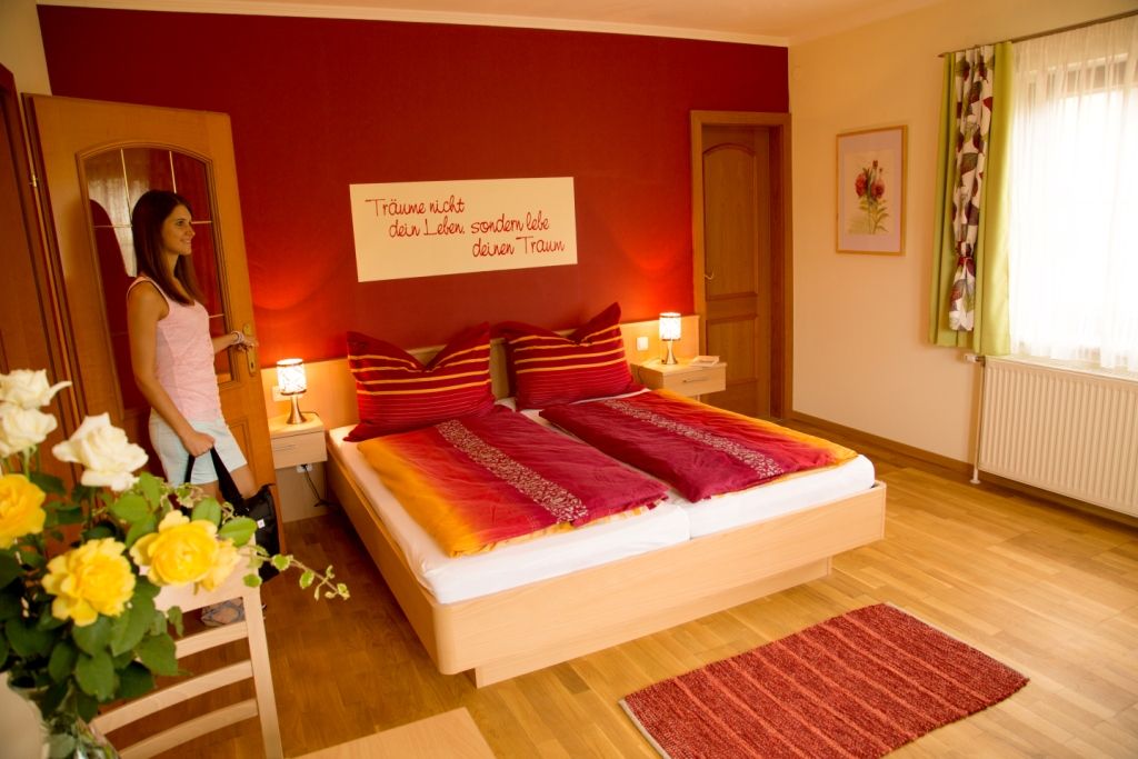 Ein gemütliches Schlafzimmer mit roter Wand, Doppelbett und Blumen. Eine Frau betritt den Raum.