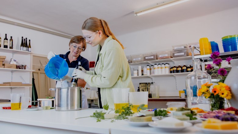 Zwei Personen arbeiten in einer Werkstatt an einem Topf mit Fl&uuml;ssigkeiten und Kr&auml;utern auf einem Tisch. Sie produzieren Seifen.