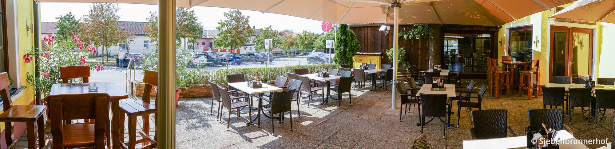 Ein sonniger Gastgarten mit leeren Tischen und Stühlen, umgeben von Pflanzen und einem großen Sonnenschirm.