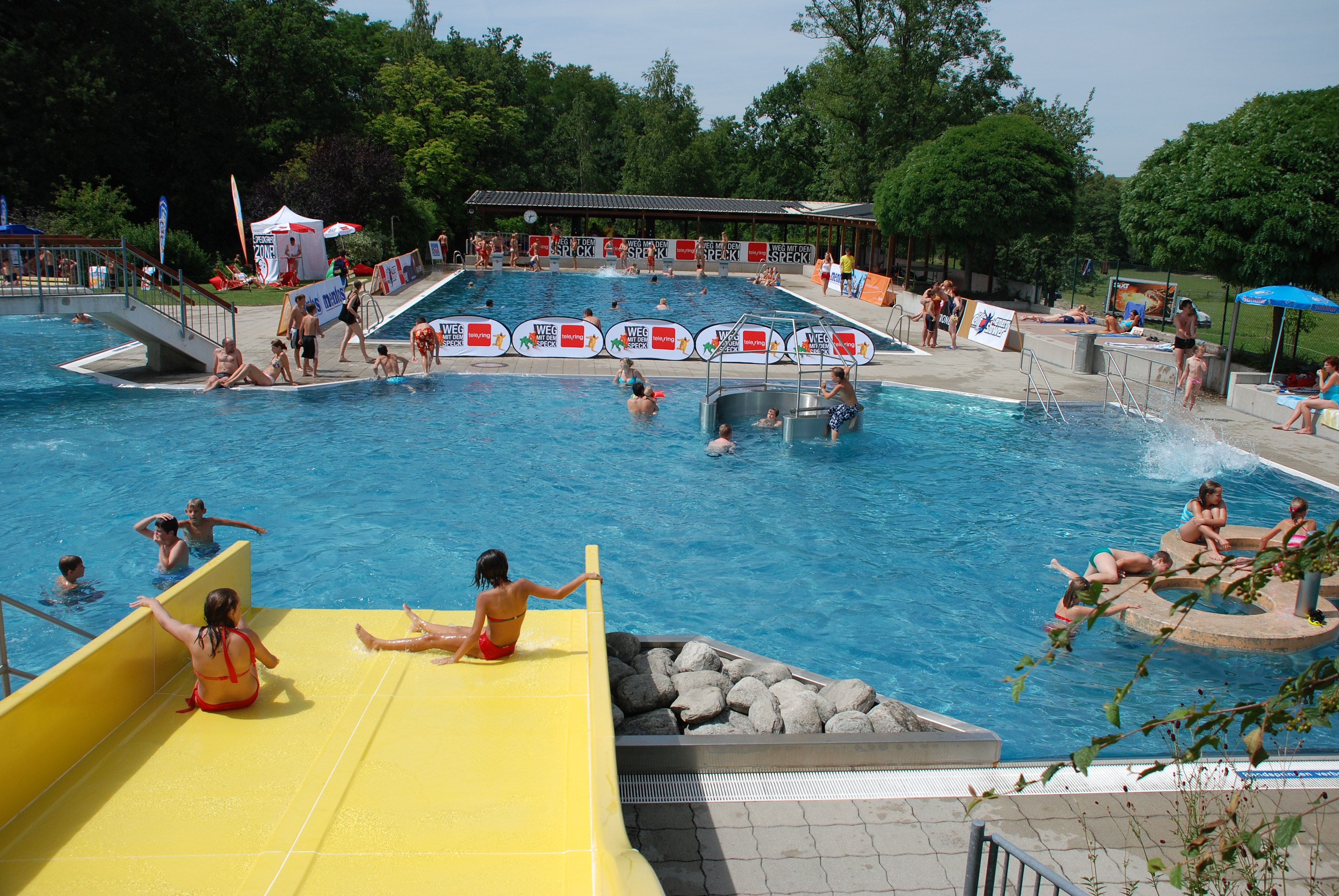 Freibad mit gelber Rutsche, Schwimmern und Liegewiese.