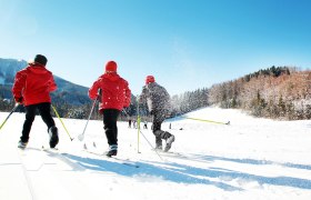 Drei Personen beim Langlaufen auf einer schneebedeckten Loipe unter blauem Himmel.