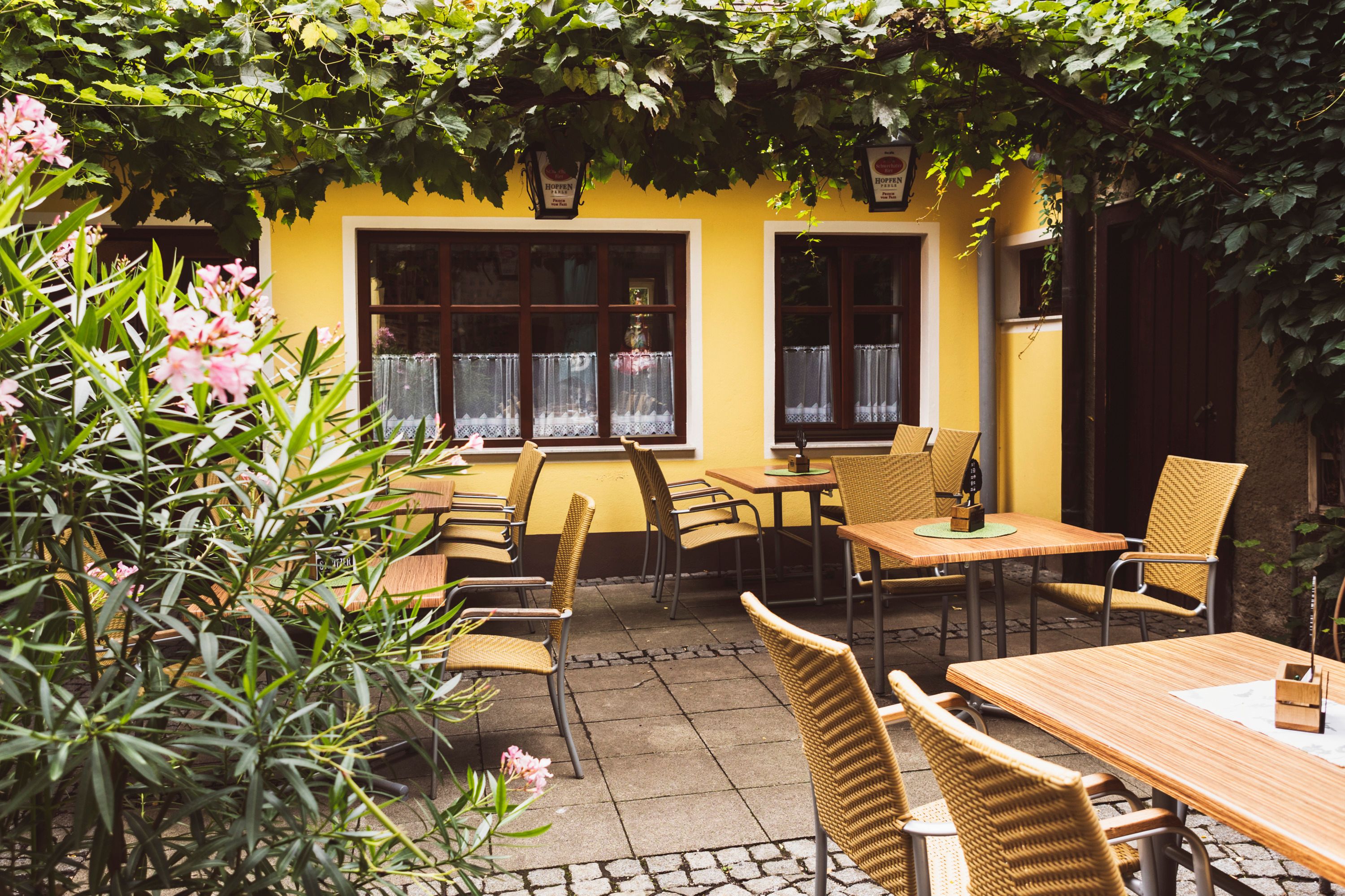 Gemütlicher Gastgarten eines Wirtshauses mit gelber Fassade, Holztischen und Korbstühlen, umgeben von Pflanzen und Weinreben.