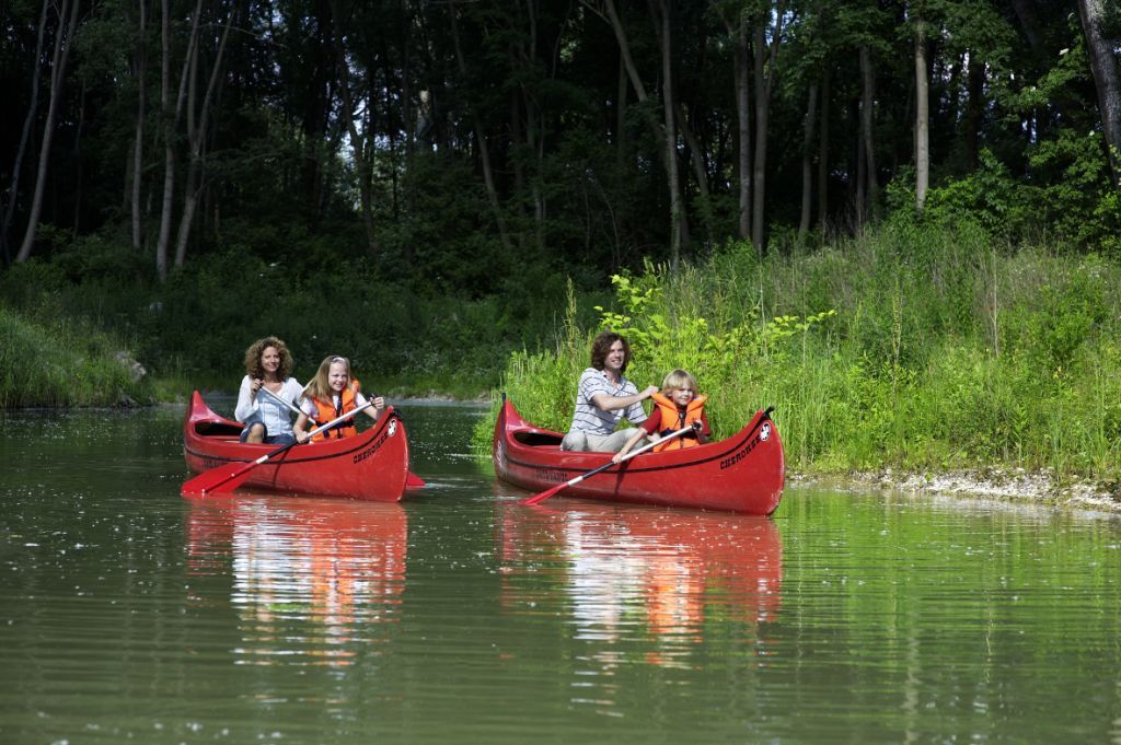 Zwei rote Kanus mit Erwachsenen und Kindern auf einem ruhigen Fluss, umgeben von grüner Vegetation.