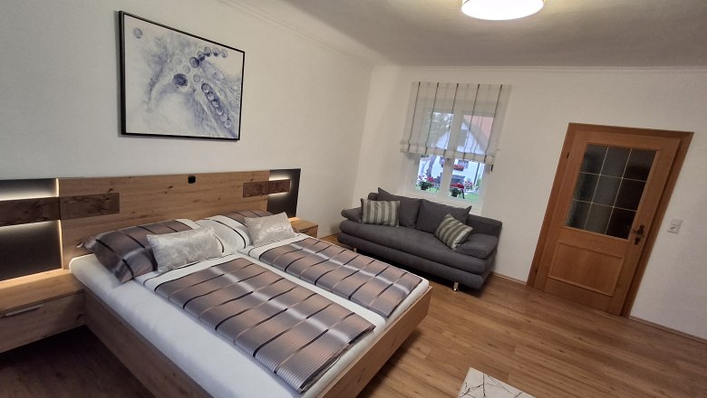 Modernes Schlafzimmer mit Doppelbett, grauem Sofa, Holzfu&szlig;boden und abstraktem Kunstwerk an der Wand.