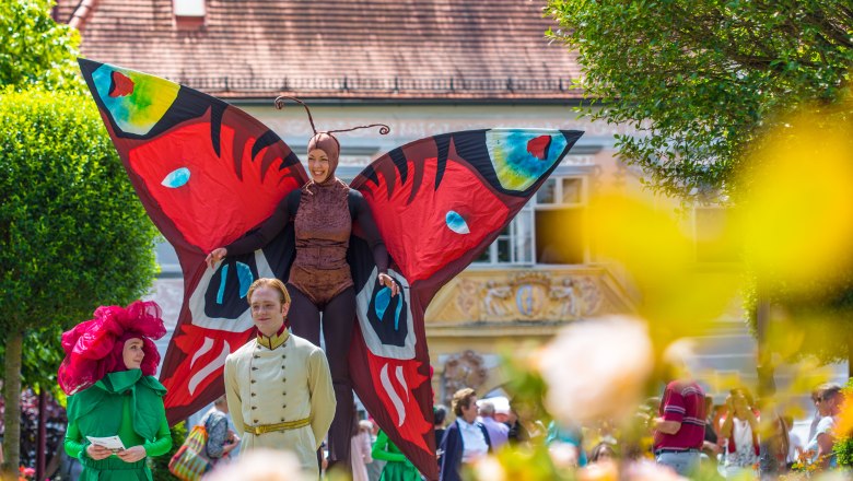Bunt verkleidete Menschen beim Rosenfest, eine Frau ist auf Stelzen als gro&szlig;er bunter Schmetterling verkleidet