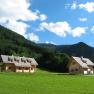 Zwei Holzh&auml;user auf einer gr&uuml;nen Wiese vor bewaldeten Bergen und blauem Himmel.