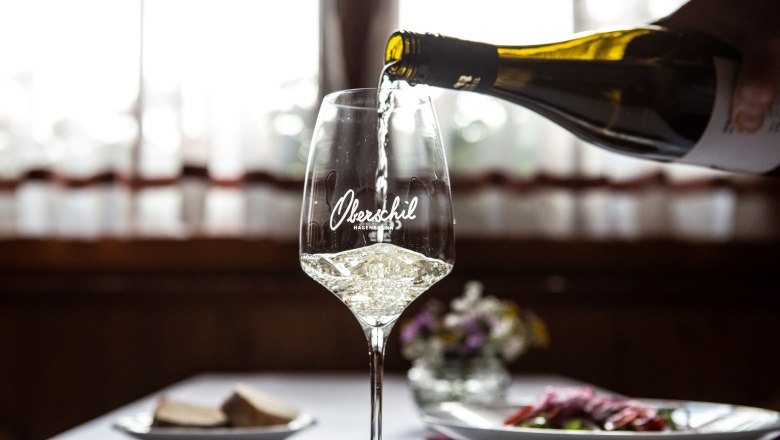 Ein Glas Wei&szlig;wein wird eingeschenkt, im Hintergrund ein gedeckter Tisch mit Blumen.