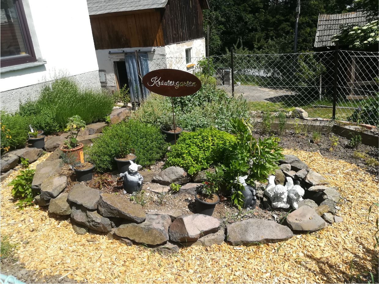 Ein Kräutergarten mit Steinumrandung, Pflanzen und Dekorationen, darunter Gartenfiguren. Ein Schild mit der Aufschrift 'Kräutergarten' ist sichtbar.
