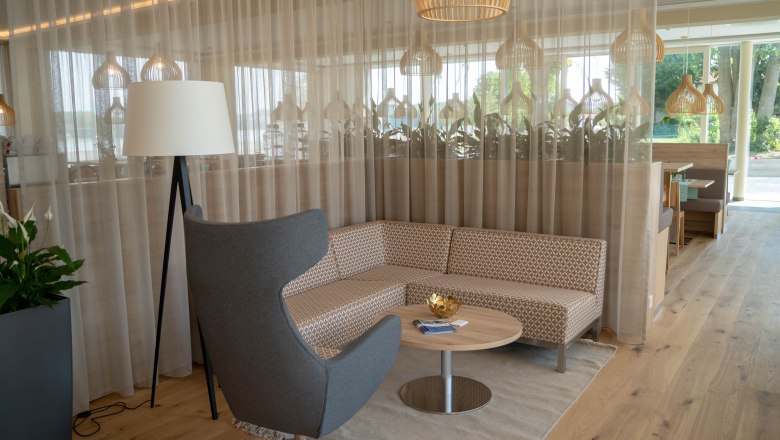Moderne Sitzecke mit grauem Sessel, gemustertem Sofa, rundem Holztisch und Stehlampe vor einem Vorhang.