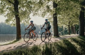 Zwei Personen, die man von vorne sieht, umgeben von Wald und Wasser, die mit ihren R&auml;dern fahren