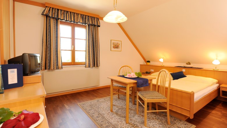 Gästezimmer im Haus Krappel, © Gästehaus Krappel Gemütliches Gästezimmer mit Holzmöbeln, Doppelbett, kleinem Tisch und Stühlen, TV und Fenster mit Vorhängen.