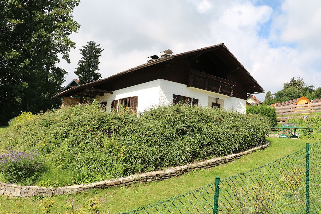 Ein weißes Haus mit Holzdach und Garten, umgeben von Büschen und Bäumen, unter einem bewölkten Himmel.