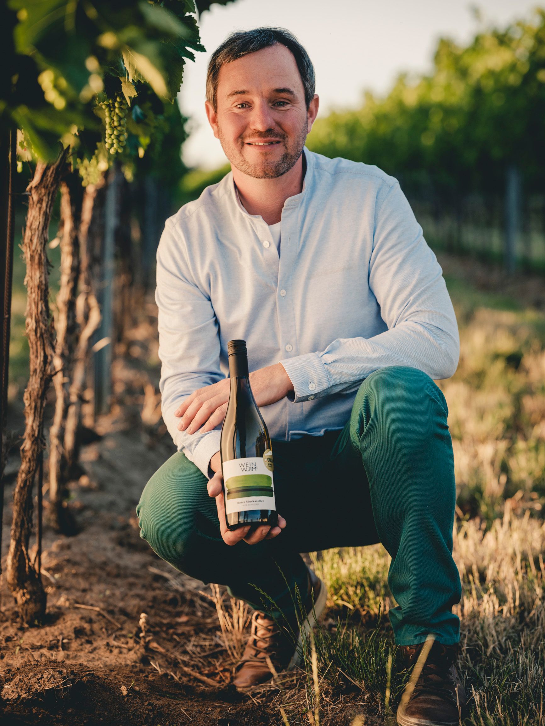 Ein Mann kniet in einem Weinberg und hält eine Weinflasche in der Hand.