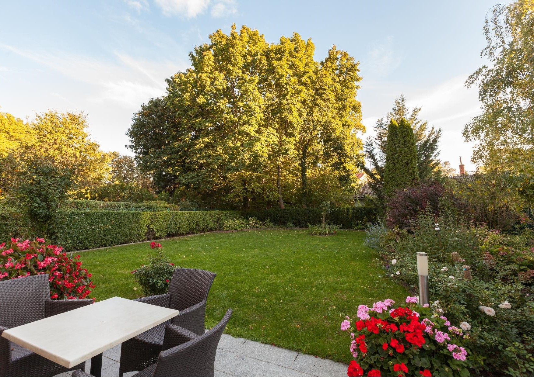 Ein gepflegter Garten mit Rasen, Blumenbeeten und Gartenmöbeln auf einer Terrasse.